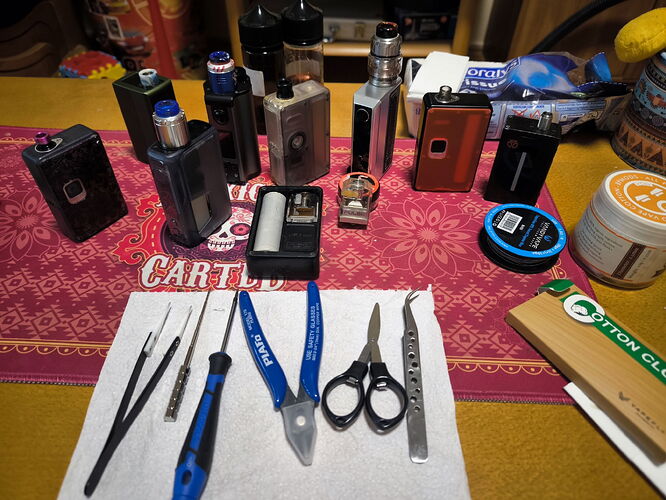 Egy asztalon különféle elektromos cigaretta eszközök, füstölők, csipeszek, olló és más kiegészítők vannak elrendezve. (Feliratozva AI-val)