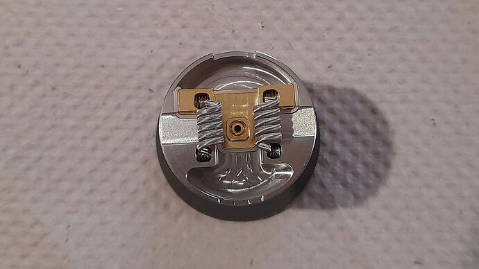 The image depicts a metal-based, dual-coil electronic device, likely a vape coil build. (Subtitled by AI)