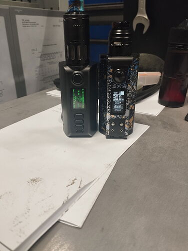 Két fekete elektromos cigaretta készülék áll egy asztalon, háttérben papírokkal és egy kulccsal. (Feliratozva AI-val)