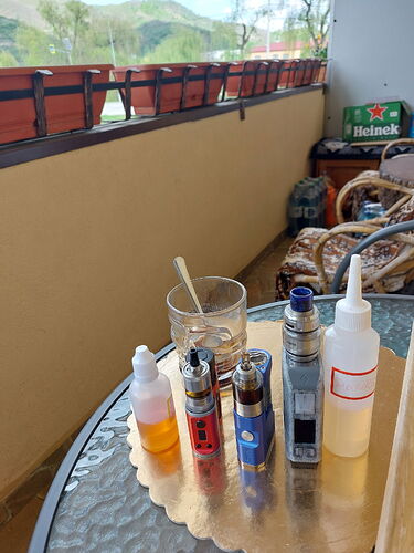 Sur la table en verre, différentes cigarettes électroniques et liquides, ainsi qu'un verre à moitié plein, se trouvent sur un balcon, avec une boîte Heineken et un paysage en arrière-plan. (Légendé par IA)