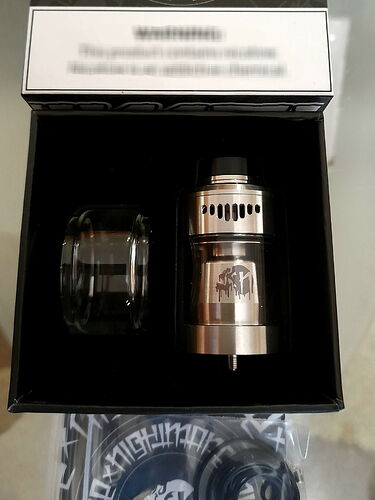 A képen egy rozsdamentes acélból készült vape tank és egy pótüveg található egy nyitott dobozban. (Feliratozva AI-val)