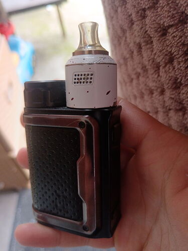 Une main tient une cigarette électronique (vape) noire avec un atomiseur blanc. (Légendé par IA)