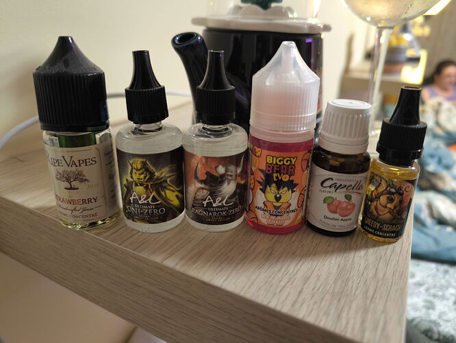 Six different e-liquid bottles lined up on a wooden shelf, with a bottle and a person visible in the blurred background. (Autogenerated by AI)