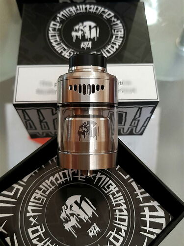 Ez az image egy fémből készült, henger alakú vape tartályt ábrázol, amely mögött egy fekete-fehér doboz és egy kör alakú minta látható. (Feliratozva AI-val)