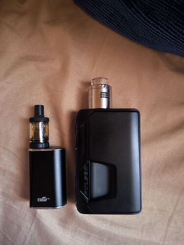 Two black electronic cigarette (vape) devices are visible on a brown surface. (Labeled by AI)