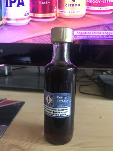 A képen egy üveg barna folyadék, rajta címkével, amely dohány kivonatra utal, egy asztalon áll, háttérben pedig egy televízió látható. (Feliratozva AI-val)