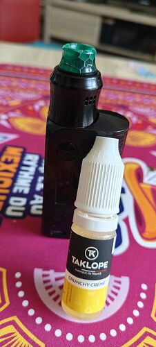 A képen egy fekete elektromos cigaretta és előtte egy TAKLOPE Crunchy Creme e-liquid tartó látható színes asztalterítőn. (Feliratozva AI-val)