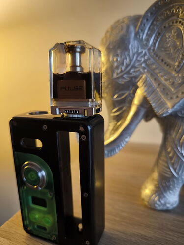 Une bouteille de parfum étiquetée 'Pulse' est placée sur le dessus d'un appareil électronique noir, s'illuminant en vert, devant une figurine d'éléphant argenté. (Légendé par IA)