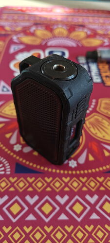 Egy fekete, hordozható hangszóró vagy elektronikai eszköz van egy színes mintás asztalterítőn. (Feliratozva AI-val)