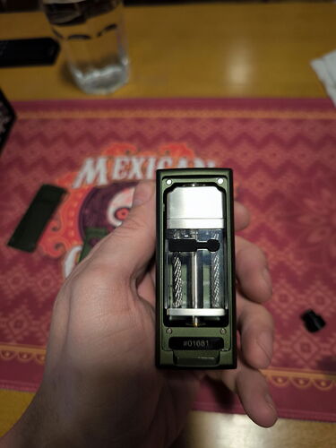The image shows a device held in hand, which is a vape box, with a transparent tank and its internal mechanism, on a Mexican-patterned mat. (Autogenerated by AI)
