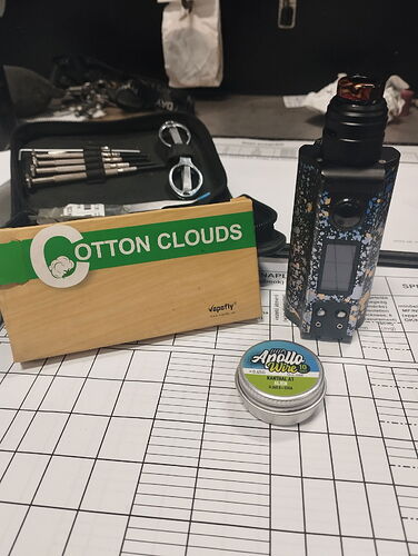 Az asztalon egy Cotton Clouds dohánycsomag, egy Apollo Breeze fémdoboz, egy elektronikus cigaretta mod és egy kis szerszámtartó látható, háttérben pedig különböző eszközök és iratok vannak. (Feliratozva AI-val)