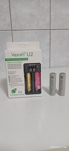 Sur l'image, la boîte d'un chargeur de batterie Vapcell U2 et deux batteries rechargeables sont visibles devant un fond de carrelage blanc. (Description générée par IA)