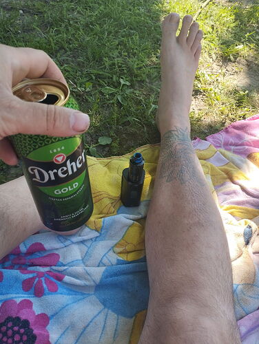 Un homme ouvre une bière Dreher Gold, les pieds nus posés sur une couverture colorée dans l'herbe. (Sous-titré par IA)