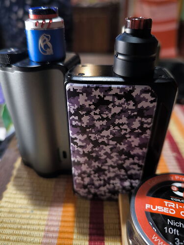 Két különböző fényű elektronikus cigaretta áll egy asztalon, egyiken kék, a másikon fekete és terepmintás burkolat van, mellettük pedig egy dohányporos doboz található. (Feliratozva AI-val)