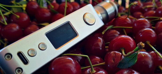 Egy ezüstszínű elektromos cigaretta fekete kijelzővel és gombokkal, cseresznyék között fekszik. (Feliratozva AI-val)