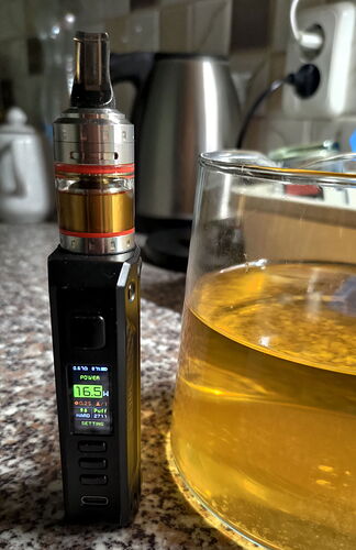 A black vape device is shown in the picture, next to it, a yellowish drink is in a transparent glass on a kitchen counter. (Captioned by AI)