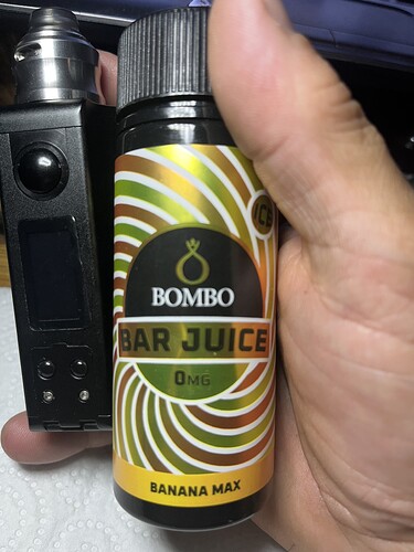 A képen egy kézben tartott e-cigaretta és egy Bombo Bar Juice nevű, banán ízesítésű, 0 mg nikotintartalmú folyadék látható. (Feliratozva AI-val)