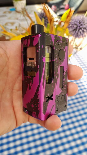 A hand holds a black and purple, splattered e-cigarette over a blue and white checkered tablecloth. (Labeled by AI)