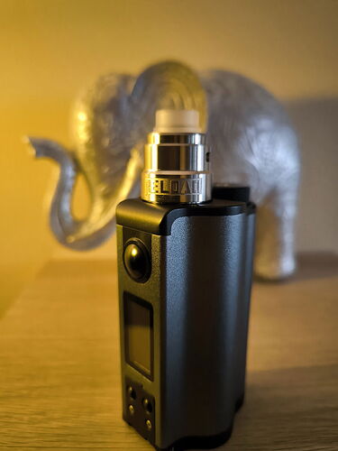 A black vape device stands on a wooden surface with a blurred silver elephant in the background. (AI-captioned)
