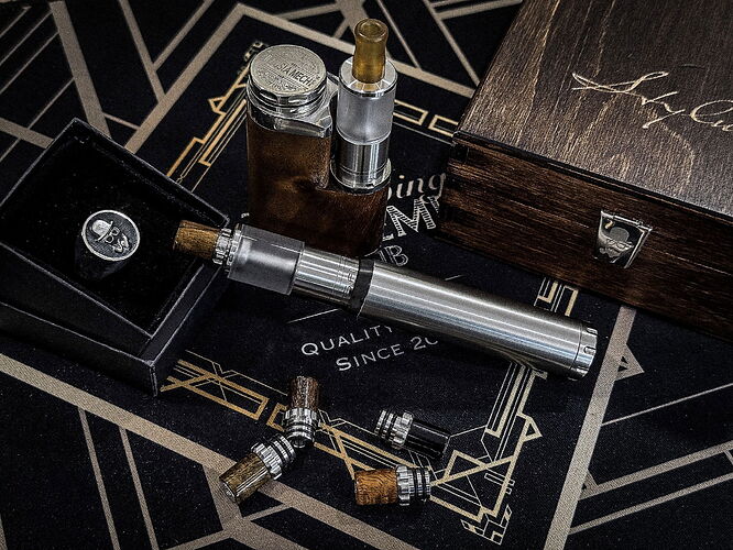 Sur la table, une cigarette électronique métallique, des ampoules de rechange, une boîte décorative en bois, ainsi qu'un cylindre métallique carré sont visibles sur un fond noir avec des motifs dorés. (Légendé par IA)
