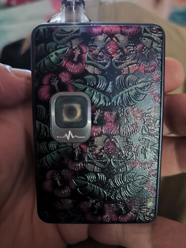 Un petit appareil cubique tenu dans une main, décoré d'un motif de feuilles sur un fond sombre, avec un petit écran et un symbole ressemblant à une forme d'onde. (Légendé par IA)