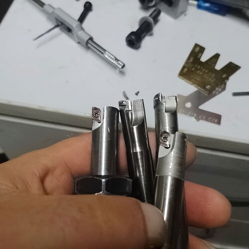 Une main tenant trois outils en métal, posés sur un établi, avec diverses pièces techniques et outils visibles autour. (Légendé par l'IA)