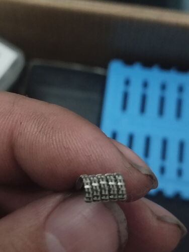 Le doigt d'une main tient un petit objet ressemblant à une chaîne métallique, avec un support en plastique bleu visible en arrière-plan. (Étiqueté par l'IA)