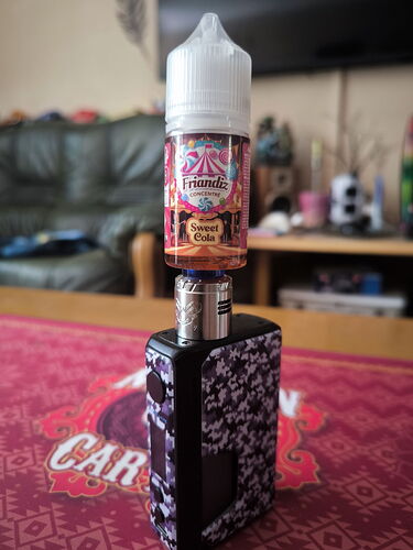 A képen egy fekete-kék mintás vape eszköz látható, amelyre egy Sweet Cola ízű, 30 ml-es e-cigaretta folyadék palack van felszerelve egy asztalon. (Feliratozva AI-val)