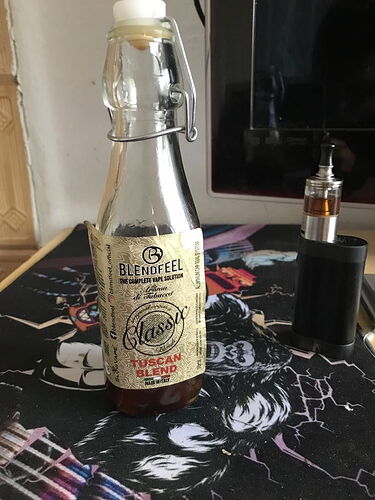 Az asztalon egy BLENDFEEL márkájú, "Classic Tuscan Blend" ízű e-liquid üveg és egy fekete dobozos e-cigaretta látható. (Feliratozva AI-val)