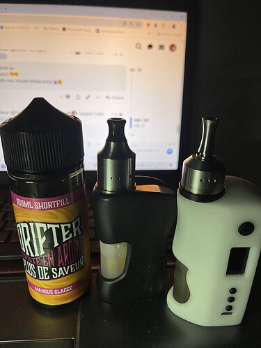 Sur la table, deux dispositifs de vapotage électroniques et une bouteille de marque "DRIFTER" contenant du e-liquide sont posés, un écran d'ordinateur est visible en arrière-plan. (Légendé par IA)