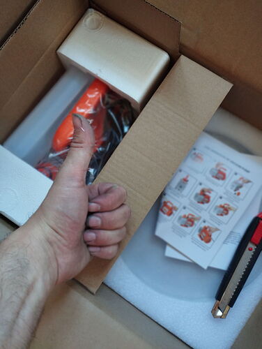 Une main fait un pouce levé au-dessus d'une boîte ouverte contenant un nouvel outil ou une pièce de machine de couleur orange, ainsi qu'un manuel d'utilisation et un cutter. (Légendé par IA)