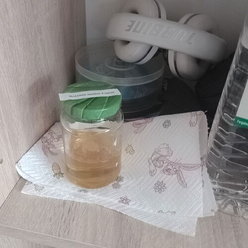 Une tasse en verre avec du thé sur une serviette en papier, avec un casque blanc et un conteneur refermable en plastique derrière, sur une étagère. (Légendé par IA)