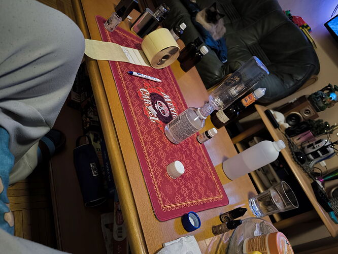 L'image montre une table avec diverses bouteilles, une bouteille d'eau, du ruban adhésif, un stylo, et un chien assis à l'arrière-plan. (Sous-titré par IA)