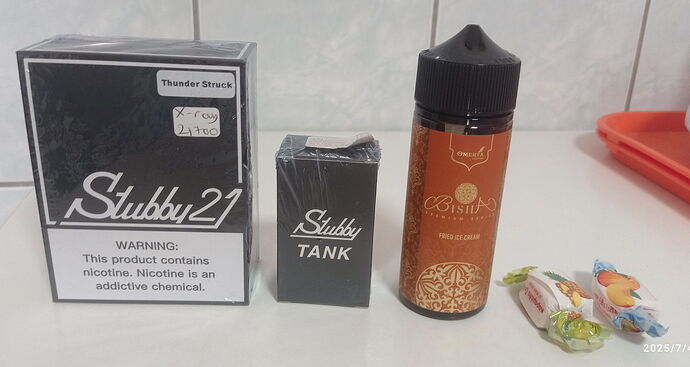 Az asztalon egy Stubby 27 doboz, egy Stubby Tank csomagolás, egy Bisita e-liquid palack és két cukorka található. (Feliratozva AI-val)