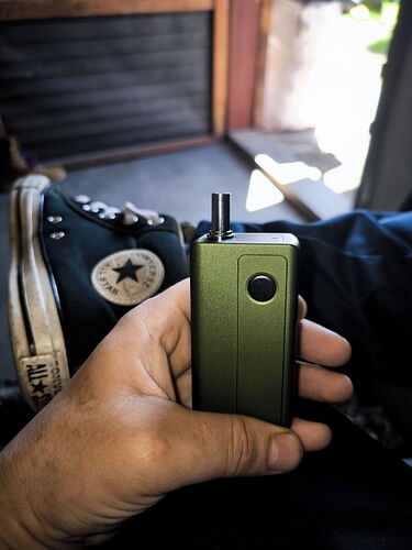 A hand holds a green e-cigarette, while a black Converse shoe is visible in the background. (Subtitled by AI)