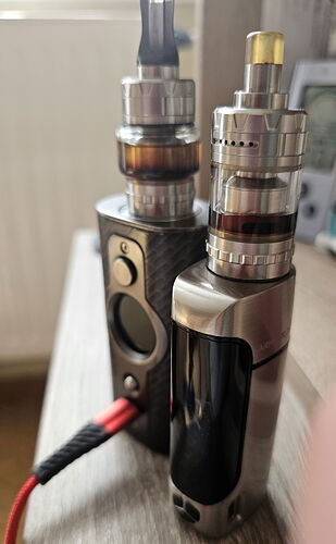 Deux dispositifs de cigarette électronique différents sont côte à côte sur une table, avec un câble rouge et noir en premier plan. (Sous-titré par IA)