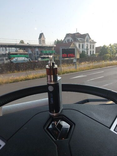 Sur le tableau de bord d'une voiture, une cigarette électronique est placée, tandis que la route et le bâtiment d'une ancienne gare sont visibles en arrière-plan. (Légendé par IA)