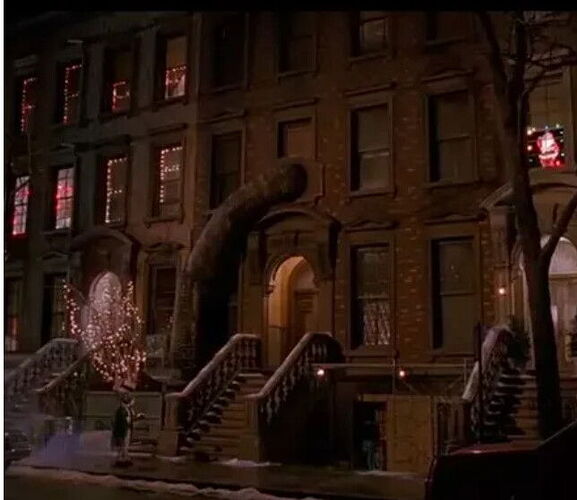 A night-time urban street view where a house's windows and a tree are decorated with Christmas lights and a huge tube or dragon-like figure clings to the building. (Labeled by AI)