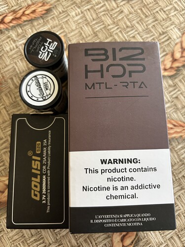 The image shows a brown B12 HOP MTL-RTA box, two small containers or caps, and a black box with gold lettering. (Subtitled by AI)