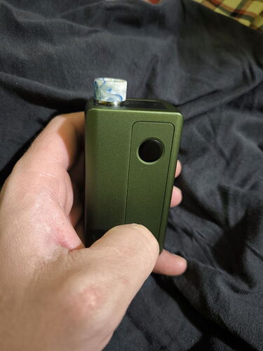 A hand holding a green, box-shaped vaporizer against a dark textile background. (AI-captioned)