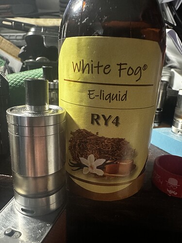 Egy White Fog márkájú RY4 jelzésű e-liquid folyadék és egy fém elektromos cigaretta tartó látható egy asztalon. (Feliratozva AI-val)