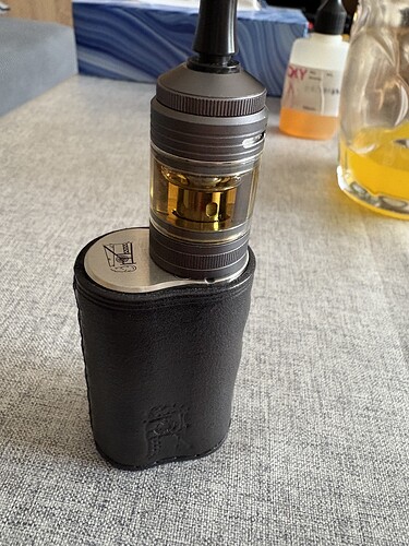 A black, textile-covered vape mod is shown, with a transparent tank and metal parts on top. (Caption by AI)