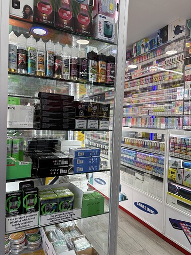 Az üzlet üvegpolcain különféle elektronikus cigaretta termékek és kiegészítők, valamint a háttérben vibrálóan világító eladótér látható. (Feliratozva AI-val)