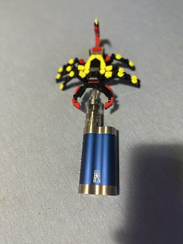L'image montre un briquet en métal bleu auquel une figurine LEGO scorpion rouge et jaune est attachée. (Légendé par IA)