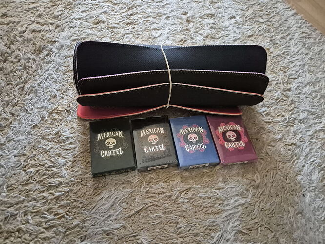 A képen egy fekete tok látható fehér szegéllyel, alatta pedig négy doboz cigaretta, amelyek mindegyike "Mexican Cartel" felirattal és egy halálfejes logóval van ellátva. (Feliratozva AI-val)