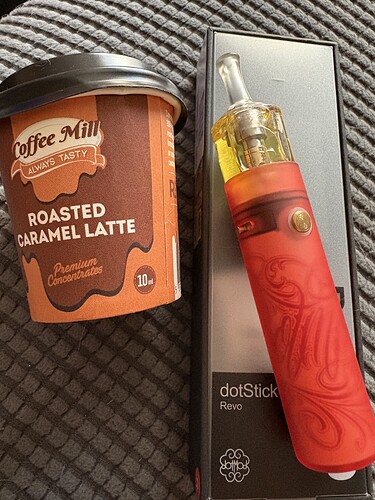 Egy sült karamellás latte pohár és egy piros mintás dotStick Revo elektromos dohányzó eszköz doboza látható egymás mellett. (Feliratozva AI-val)