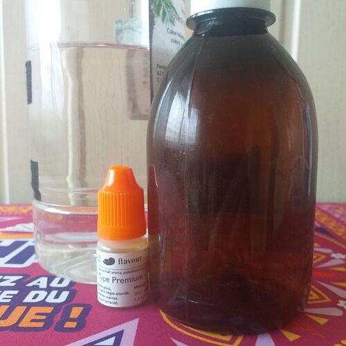 In the image, a glass bottle, a small bottle of flavoring, and a measuring cup full of water are visible on a colorful tablecloth. (AI captioned)