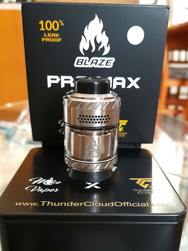 Az képen egy fényes, ezüstszínű vape atomizer látható, mely előtt egy fekete doboz áll a Blaze Promax márkajelzéssel és egy tűzszimbólummal. (Feliratozva AI-val)