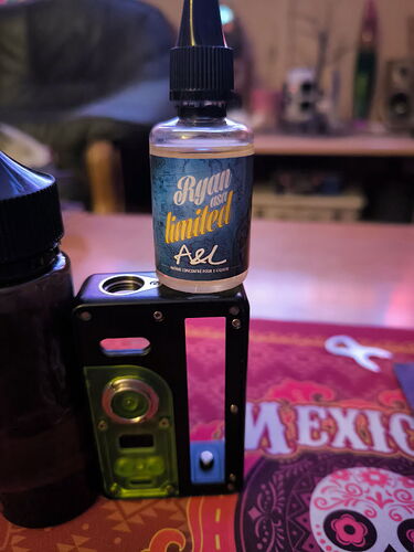 Az asztalon egy kék és zöld színű vape mod van, amely tetején egy Ryan-limited A&L folyadék üveg áll. (Feliratozva AI-val)