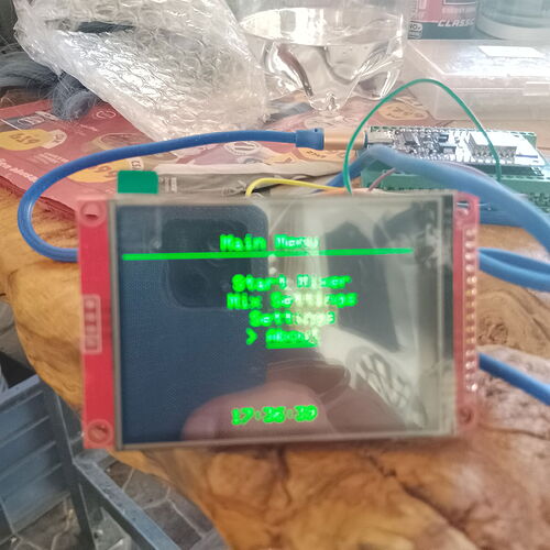 A close-up shot shows a small electronic display showing a menu in green text, connected to other electronic components and blue wires on a surface. (Caption by AI)